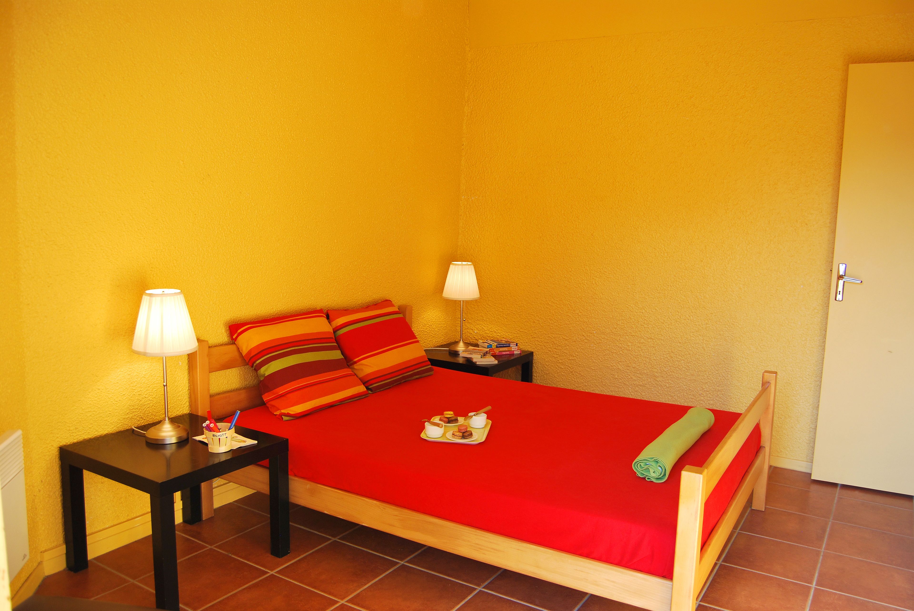 Una cama individual con una colcha roja, dos mesitas de noche con lámparas y un cabecero de madera.