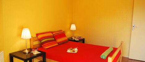 Una cama individual con una colcha roja, dos mesitas de noche con lámparas y un cabecero de madera.