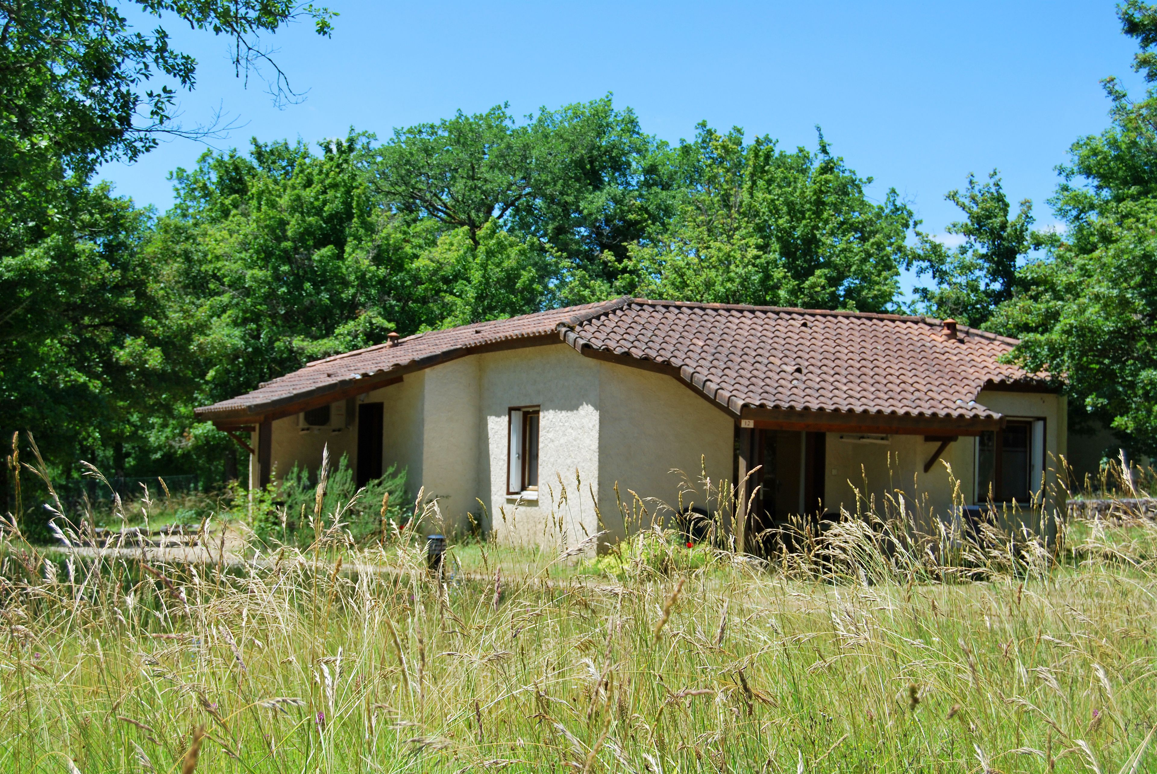 A rustic house with a tiled roof, surrounded by tall grass and trees.