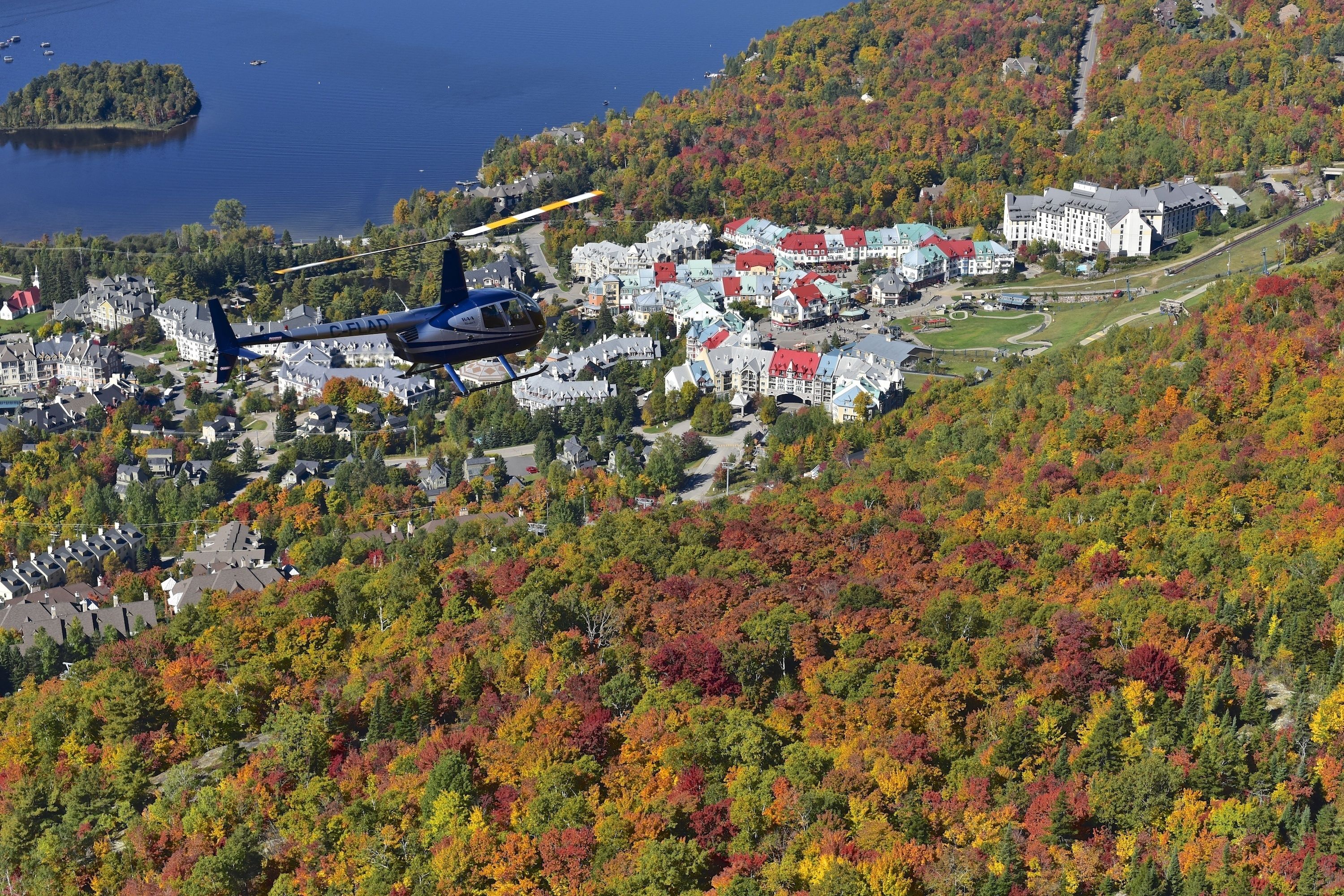 A helicopter landing on a helipad near a town with colorful autumn foliage and a lake in the background.