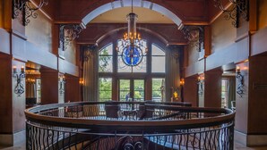A circular staircase with ornate railings, a chandelier, and large windows offering a view of greenery.