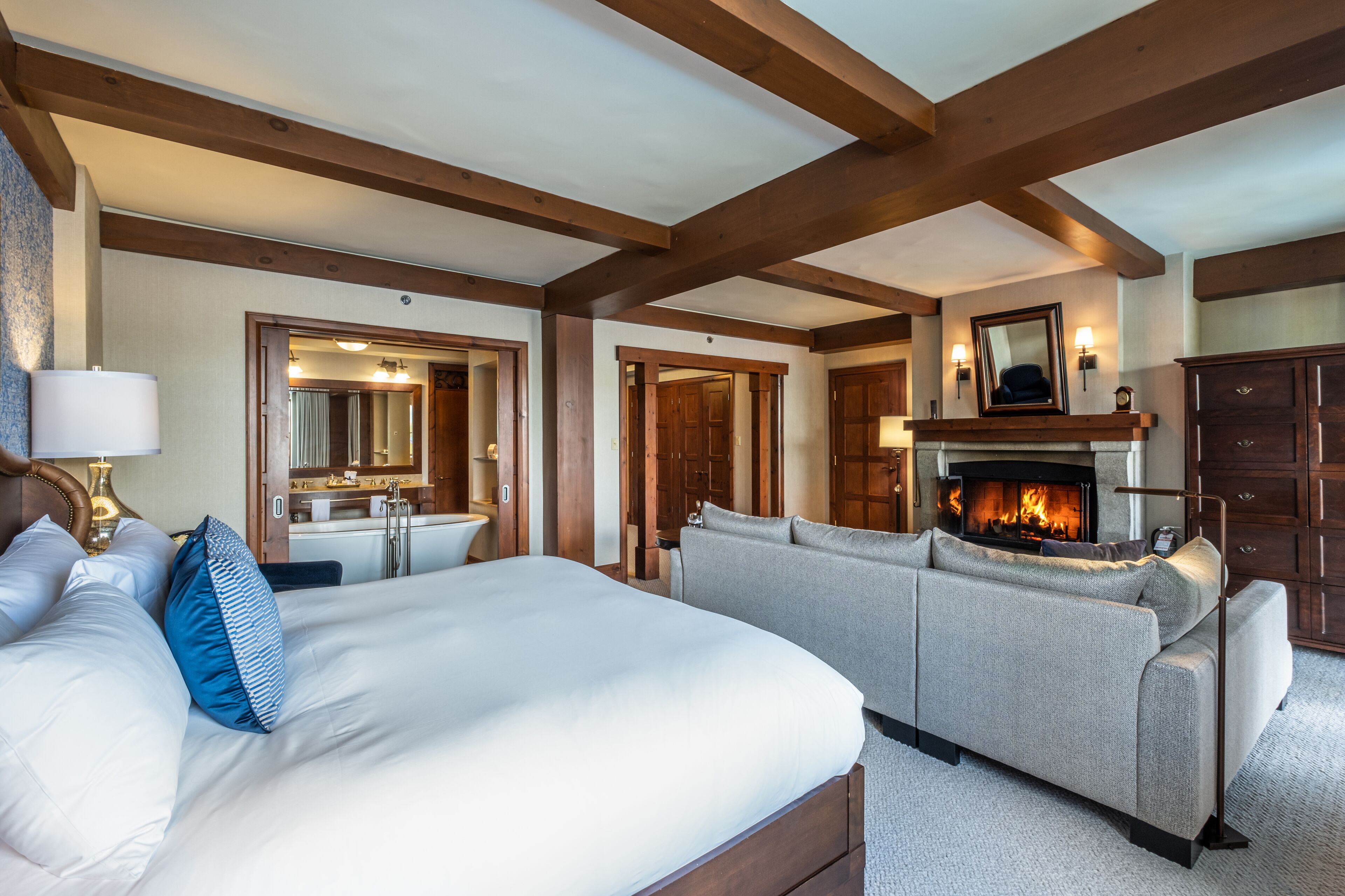 A hotel room with a large bed, a sofa, a fireplace, and wooden beams on the ceiling.
