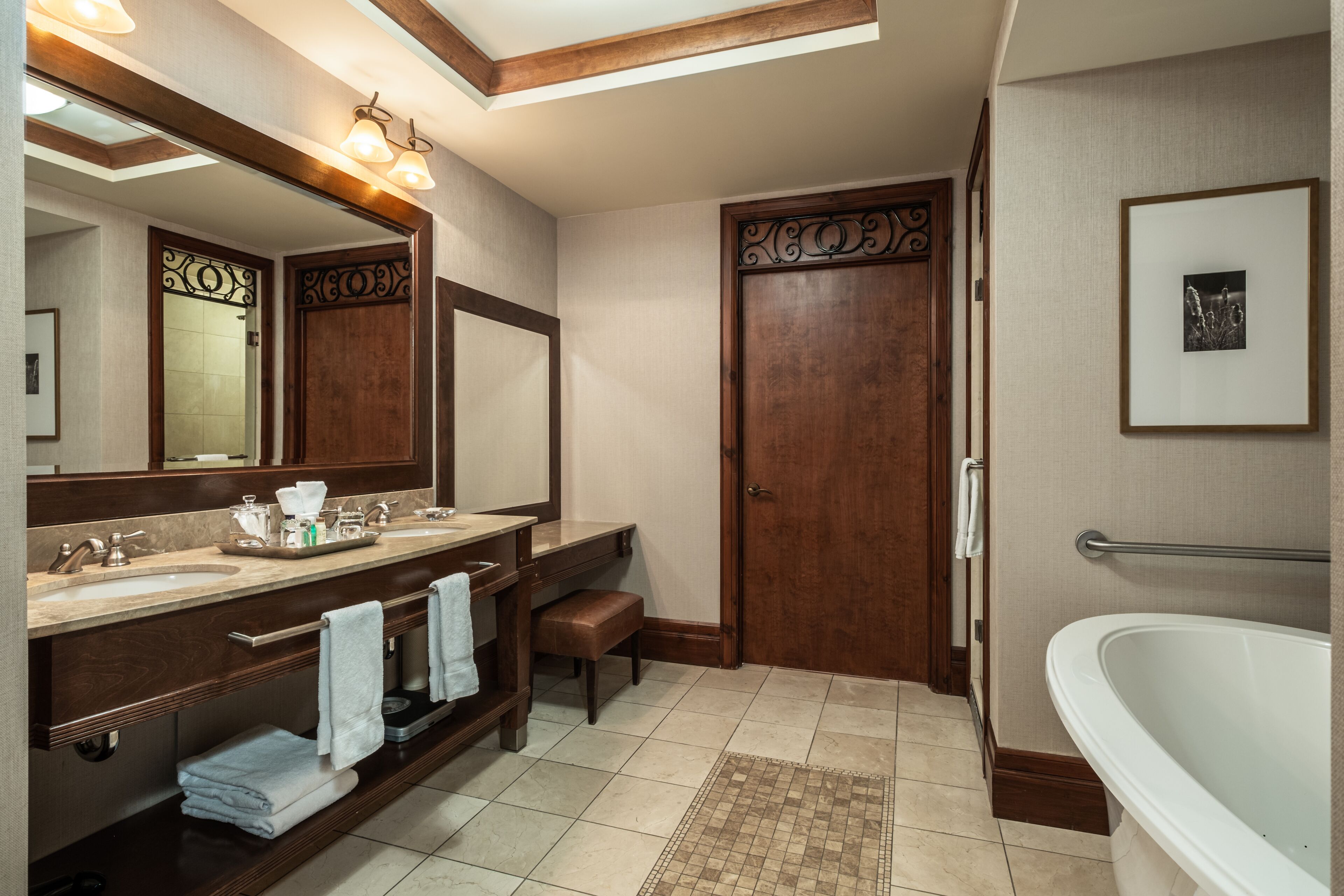 Un baño con tocador doble, un espejo grande, una puerta de madera, una bañera y un pequeño banco.