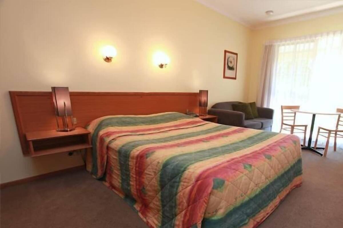 A hotel room with a bed, bedside tables, a chair, a small table, and a window with curtains.