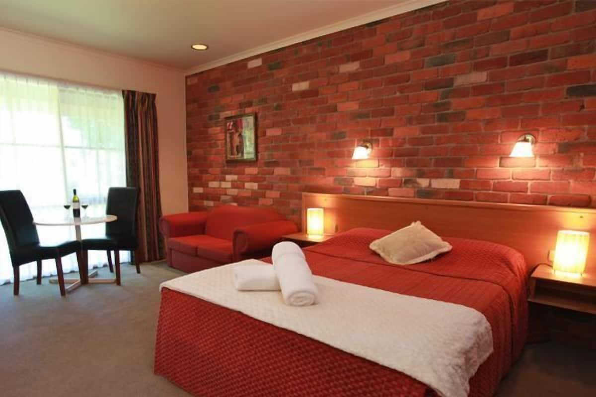 A hotel room with a brick wall, a red bed, a red sofa, a small table with a bottle, and two bedside lamps.
