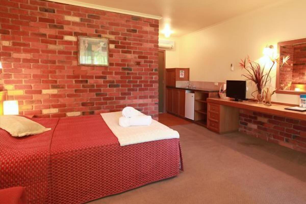 A hotel room with a brick wall, a bed with a red bedspread, a desk with a computer, and a small kitchenette.