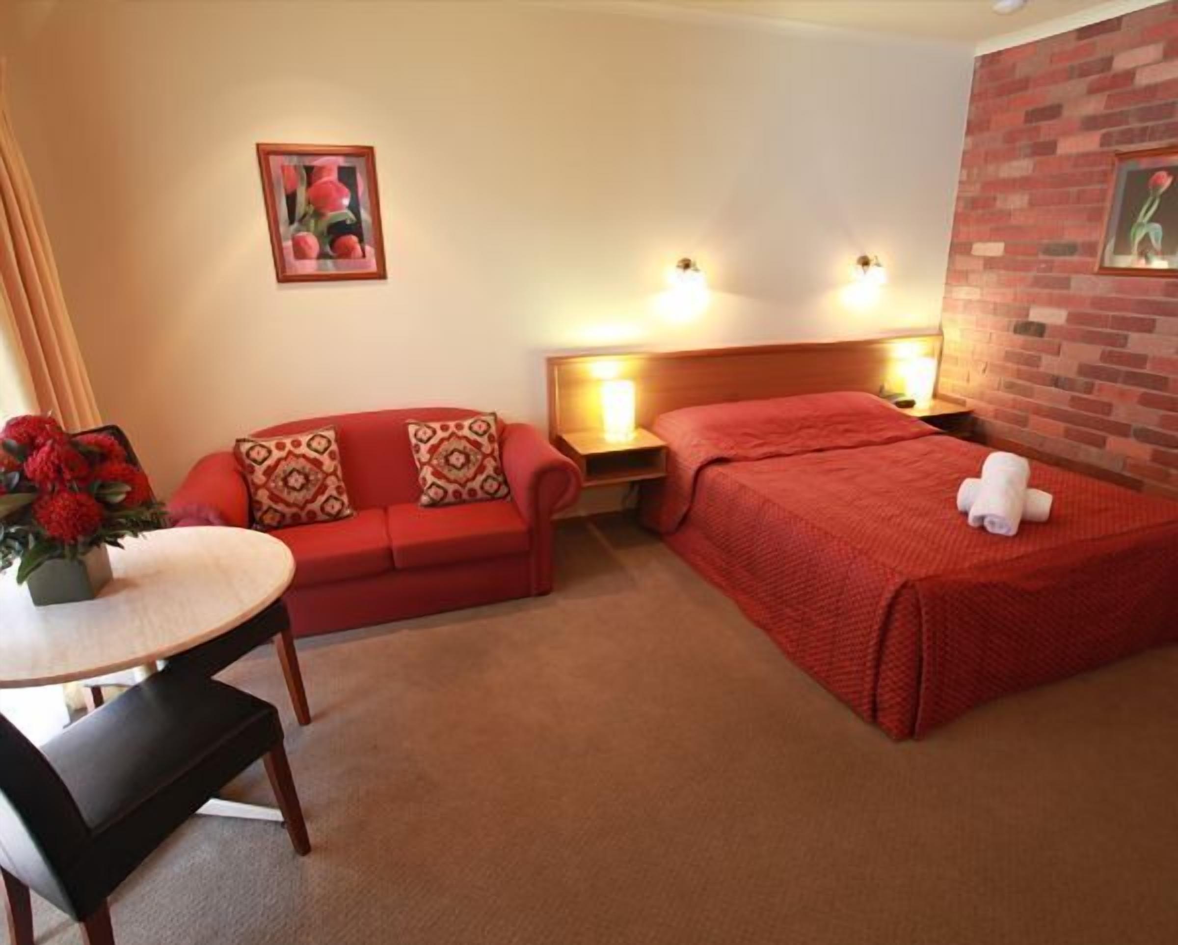 A hotel room with a red sofa, a single bed, a small table with a vase of flowers, and a brick accent wall.