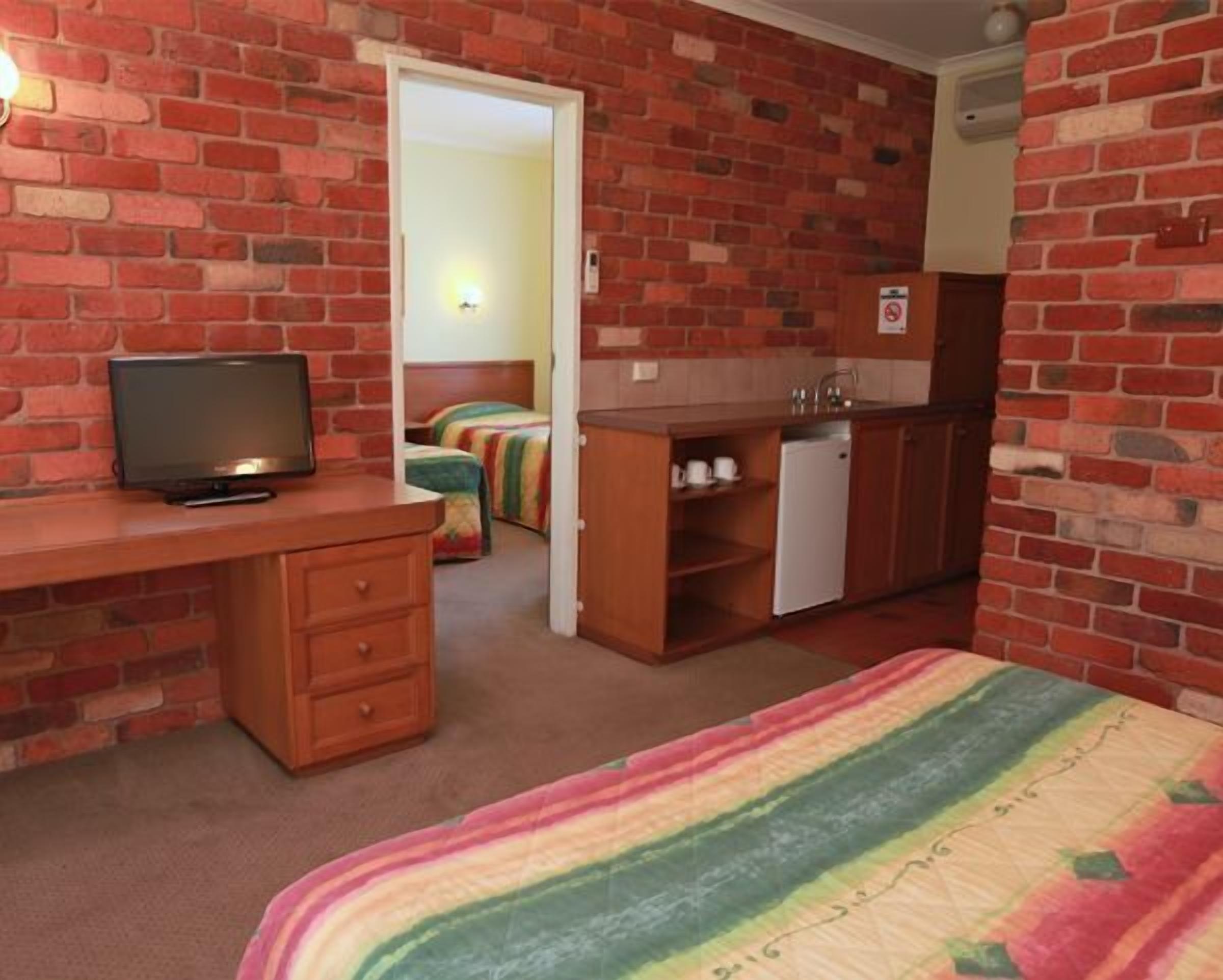 A room with a brick wall, a television on a wooden desk, a bed with a rainbow-striped blanket, a kitchenette with a refrigerator, and a door leading to another room.