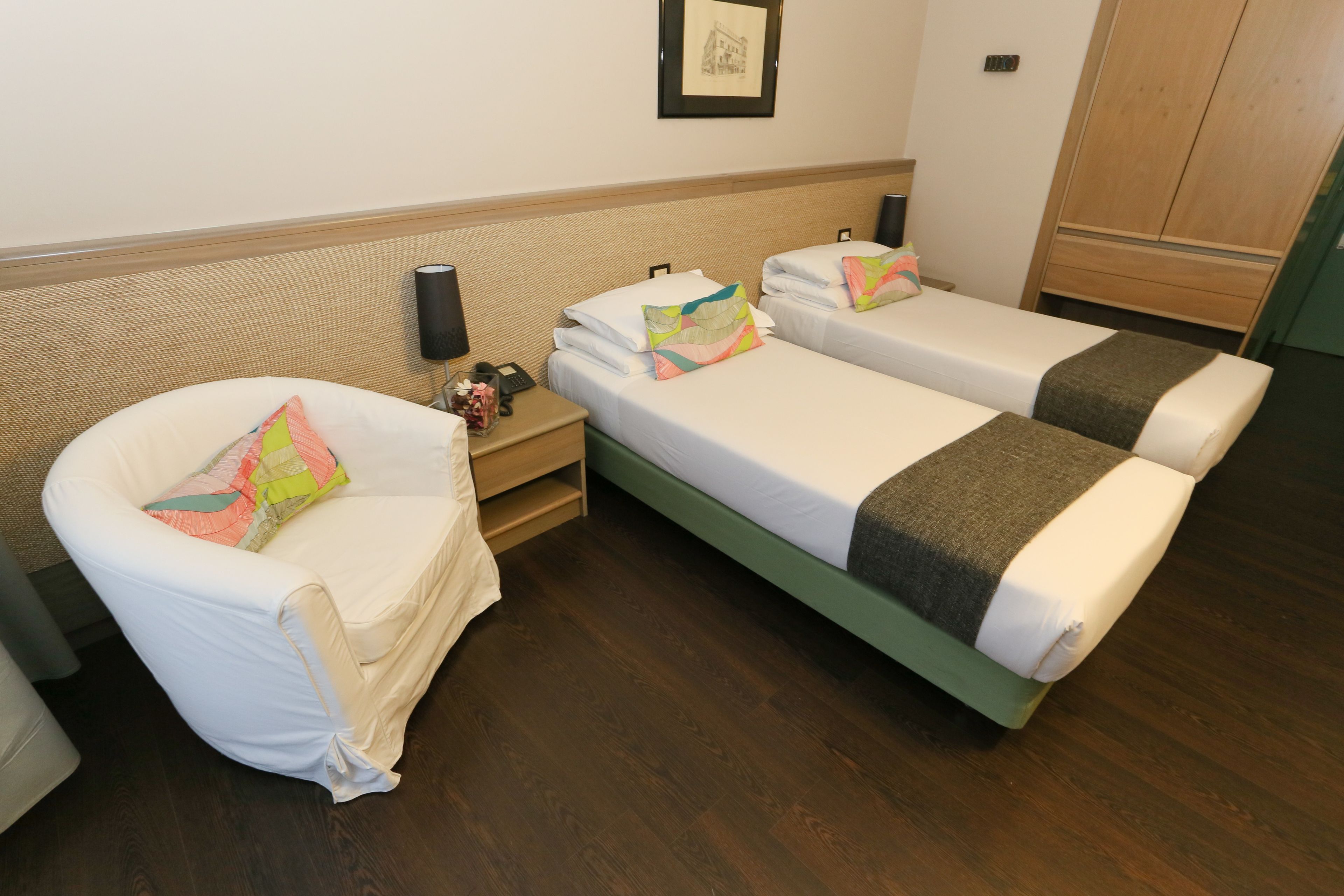 A hotel room with two single beds, a chair, a nightstand, and a wardrobe.