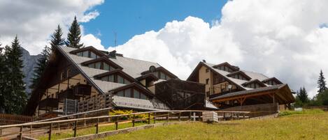 Un edificio de estilo chalet con varios niveles, balcones de madera y techo inclinado, ubicado en una ladera cubierta de hierba con montañas al fondo.
