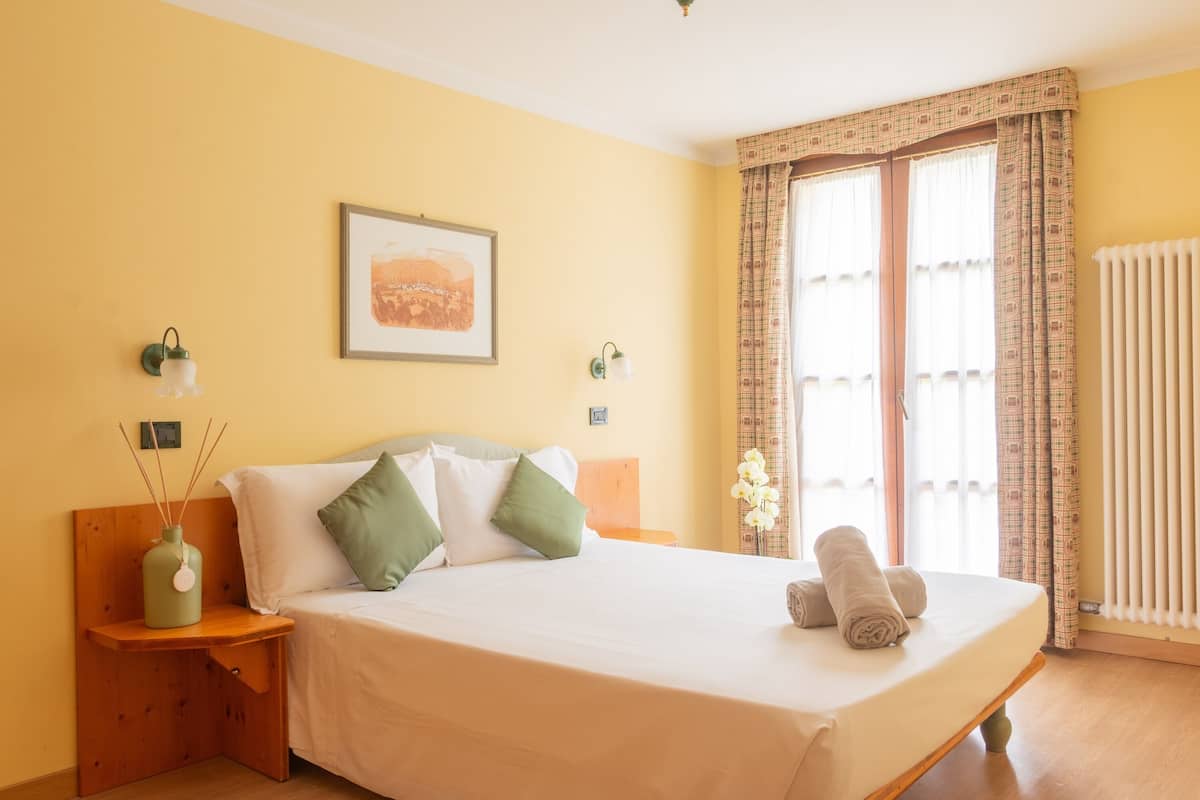 A bedroom with a bed, bedside table, wall-mounted lamp, and a window with curtains.