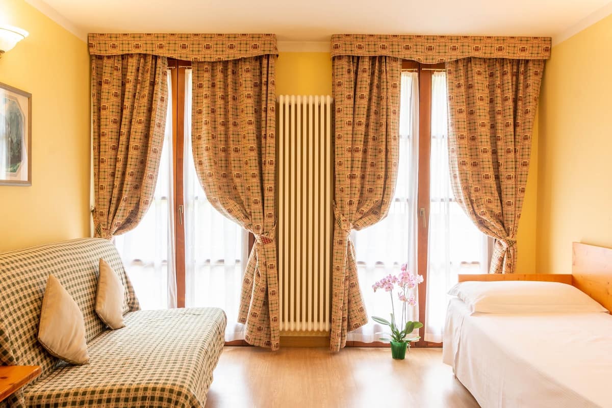 A hotel room with a checkered sofa, a bed, and a radiator between windows with curtains.