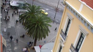 A street view with a yellow building, palm trees, and a few pedestrians. - Claridge Hotel (Athens)