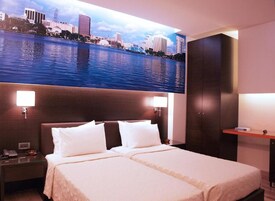 Une chambre d’hôtel avec un grand lit, deux tables de chevet, une commode, un bureau et un téléviseur fixé au mur qui diffuse des images d’un paysage urbain.