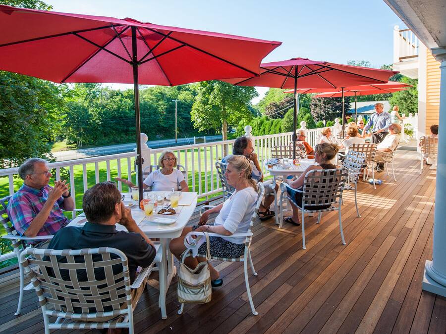 A covered outdoor deck with tables and chairs, surrounded by a white fence, overlooking a green landscape.