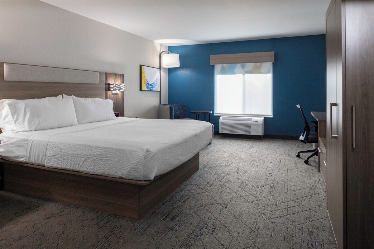 A modern hotel room with a large bed, a desk, a chair, a lamp, and a window with blinds.