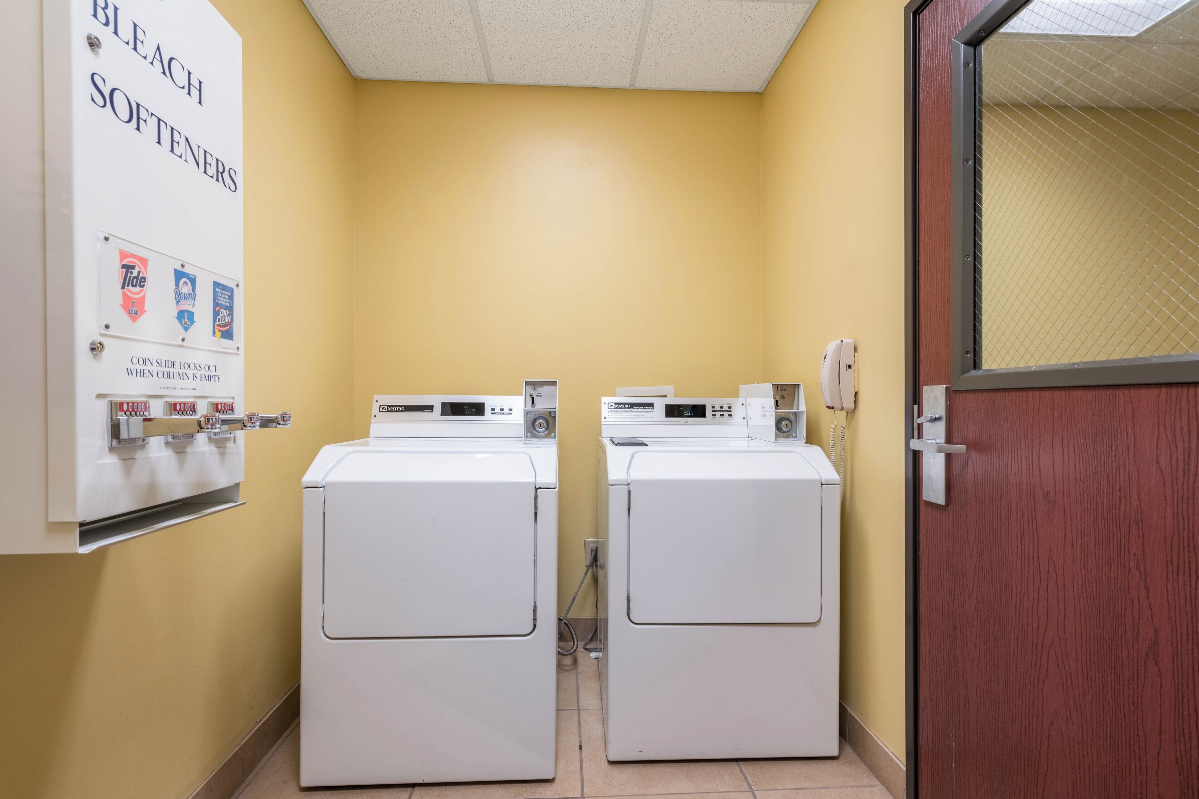 A laundry room with two washing machines, a coin slot for laundry supplies, and a door with a security lock.