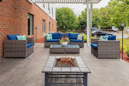 A patio area with wicker furniture, a fire pit, and a brick building in the background.