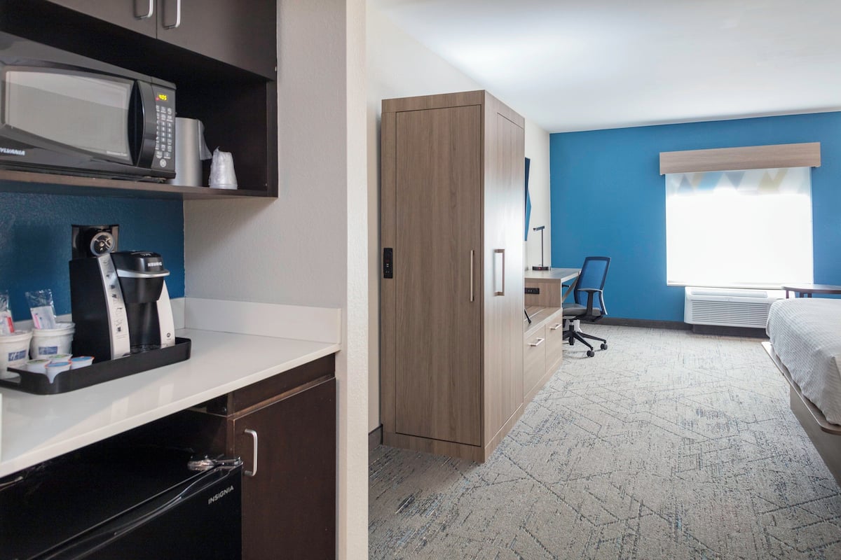 A hotel room with a kitchenette, a bed, a desk, and a window with curtains.
