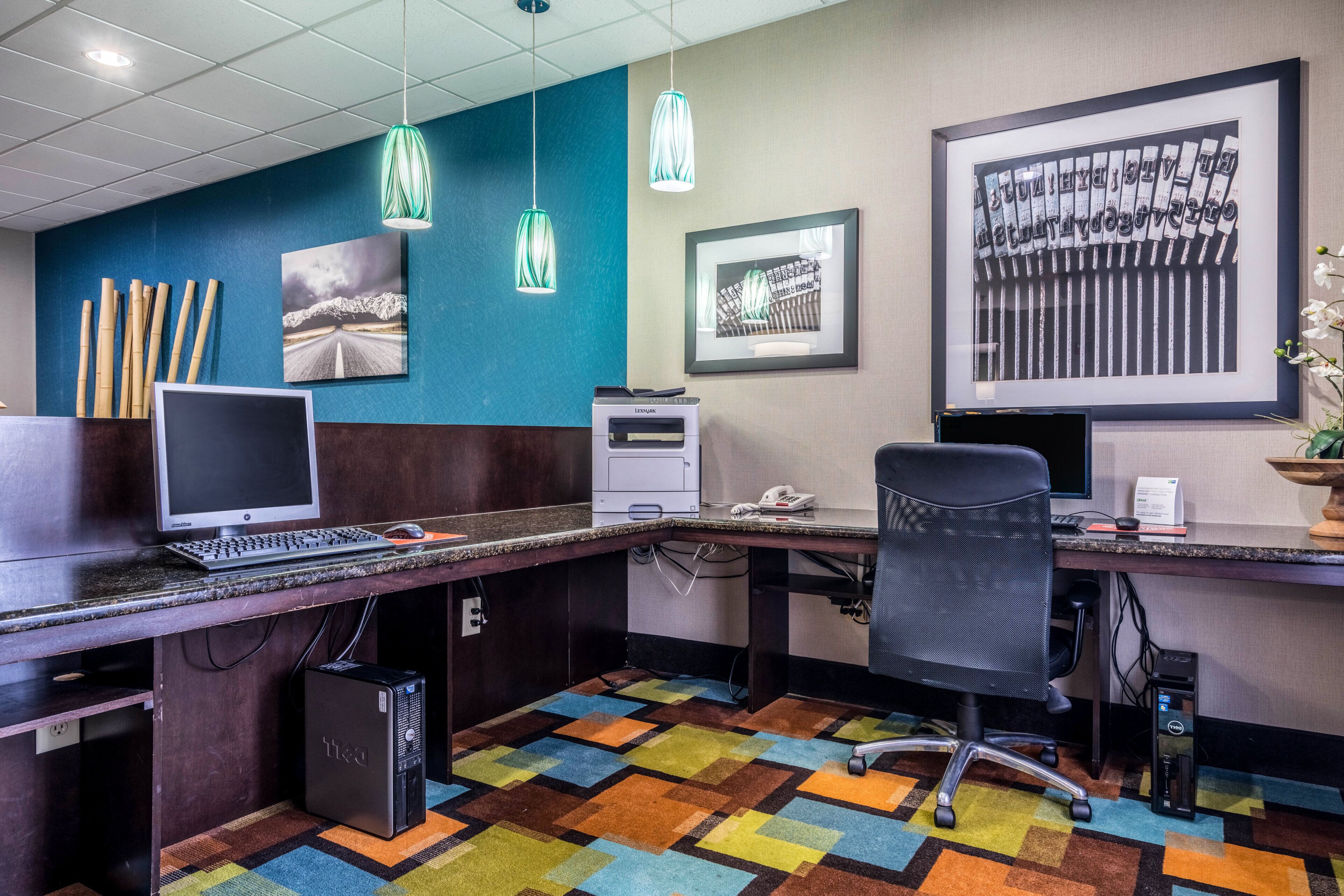 Un espace de bureau moderne avec un bureau, un ordinateur et un fauteuil de bureau. Des tableaux encadrés ornent le mur et un tapis coloré recouvre le sol.