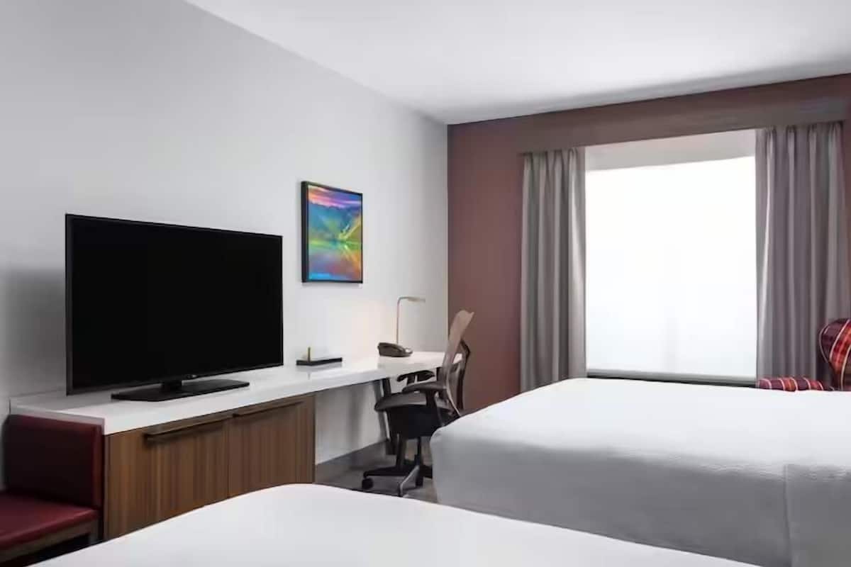 A hotel room with a flat-screen TV, a desk with a lamp, two beds, a chair, and a window with curtains.