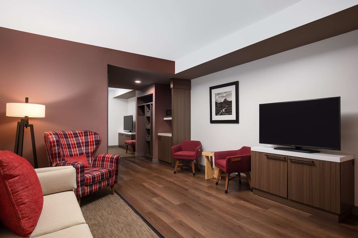 A modern living room with a sofa, armchair, and red chairs. A flat-screen TV is mounted on a wooden cabinet.