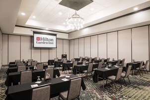 Un salón de conferencias con varias mesas y sillas, una pantalla de proyector mostrando 'Hilton Garden Inn' y un candelabro.