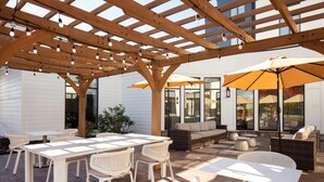 Un patio exterior con pérgola de madera, mesas, sillas y una zona de sofás con sombrillas.