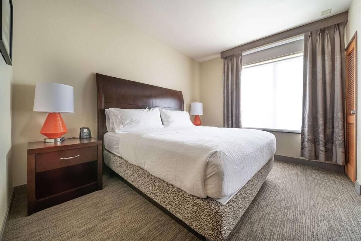 A hotel room with a large bed, two bedside tables with lamps, a window with curtains, and a carpeted floor.