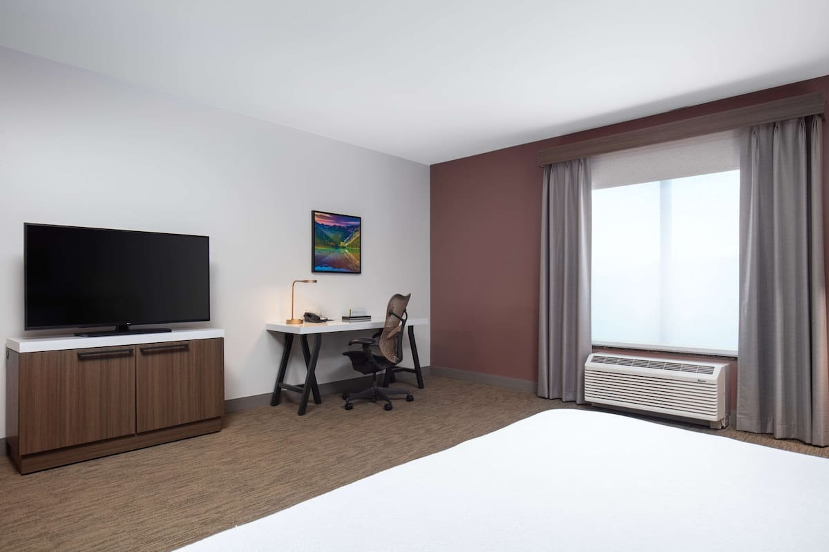 A hotel room with a flat-screen TV, a desk with a lamp, a chair, a bed, and a large window with curtains.
