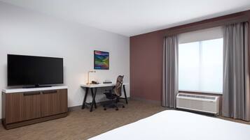 A hotel room with a flat-screen TV, a desk with a lamp, a chair, a bed, and a large window with curtains.
