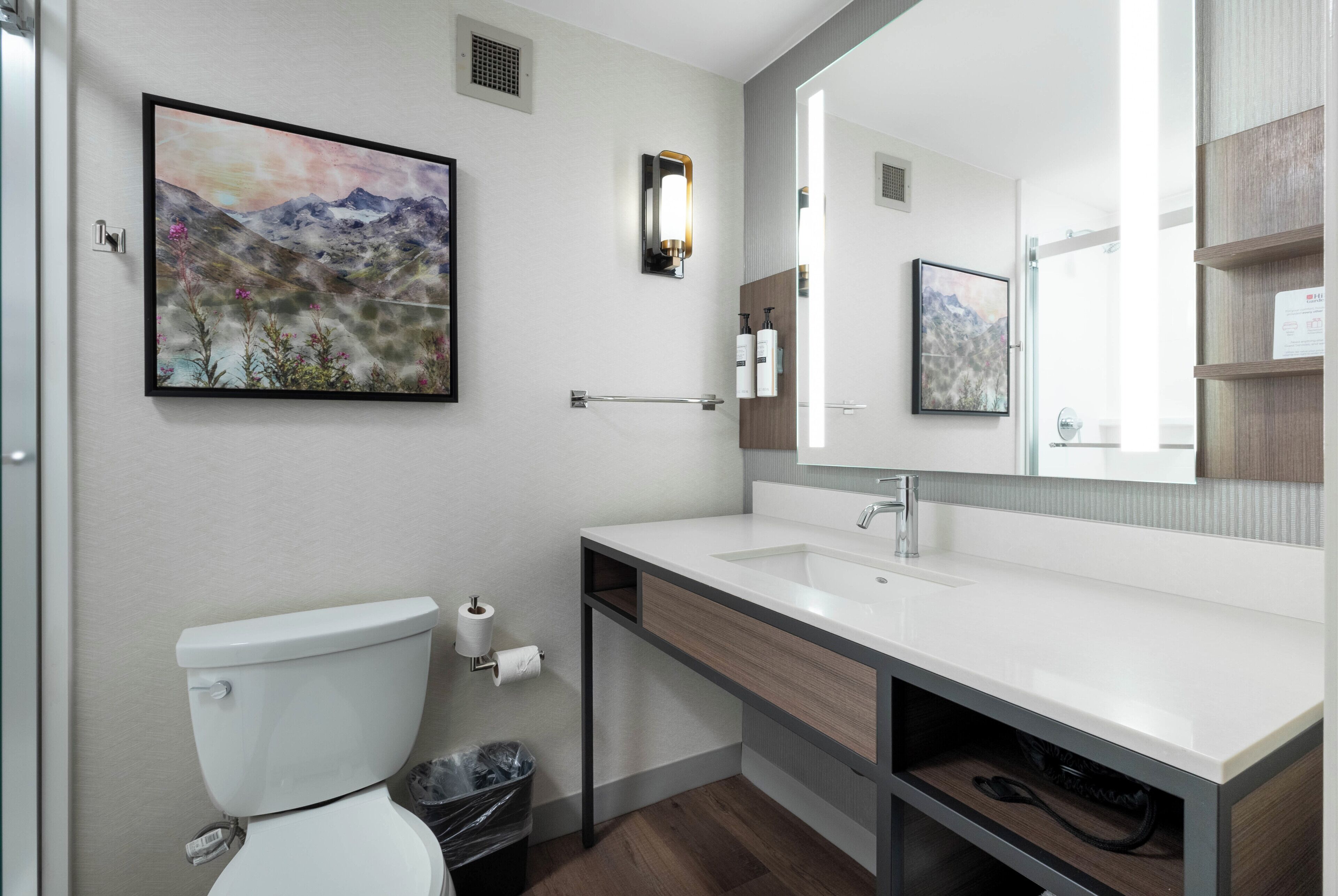 A modern bathroom with a toilet, sink, mirror, and a framed landscape painting on the wall.