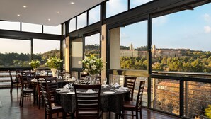 Un restaurant avec des tables rondes disposées pour le service, de grandes fenêtres offrant une vue sur un bâtiment historique et des arbres.