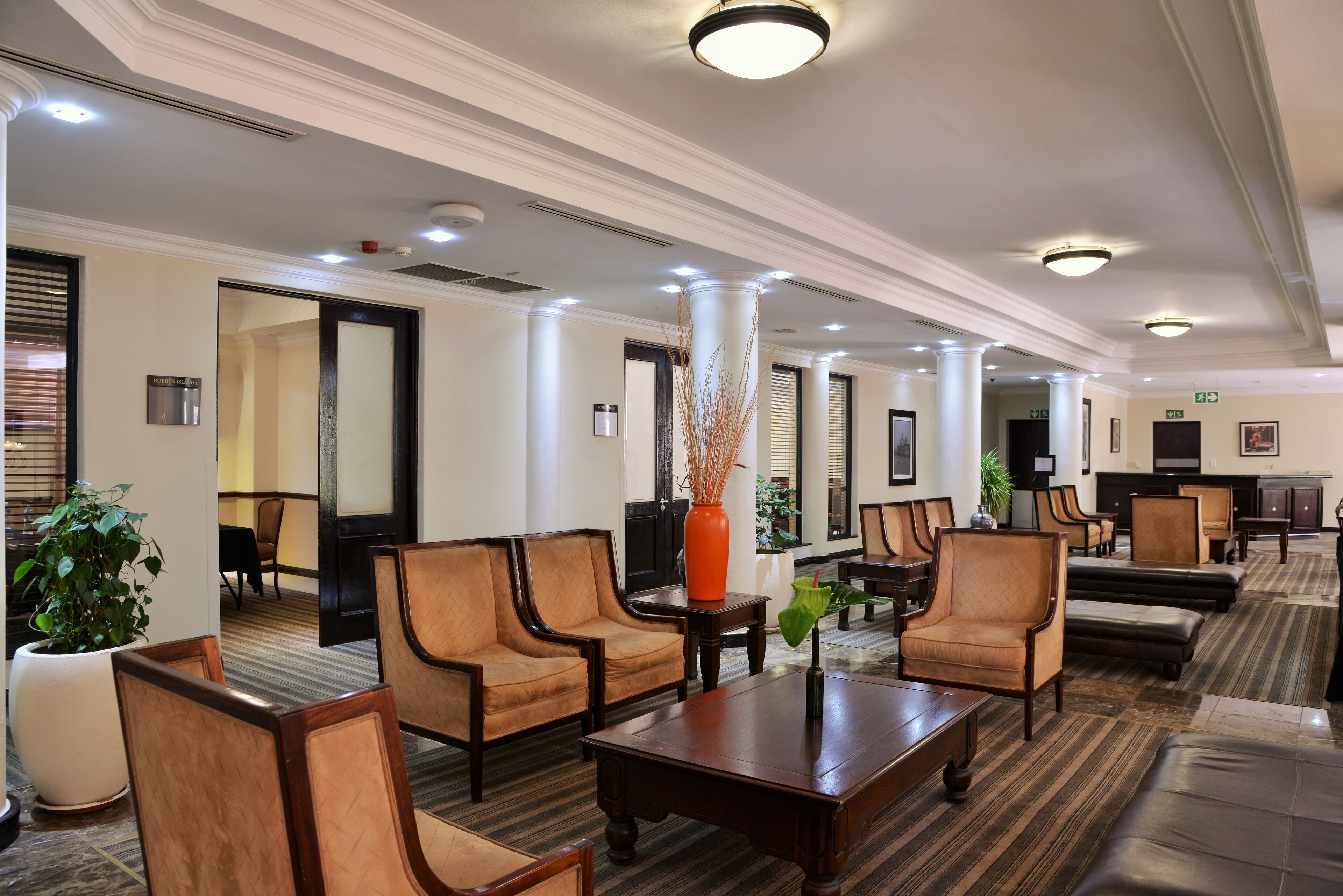 Un vestíbulo de hotel con piso alfombrado, mobiliario de madera y un jarrón grande con plantas secas.