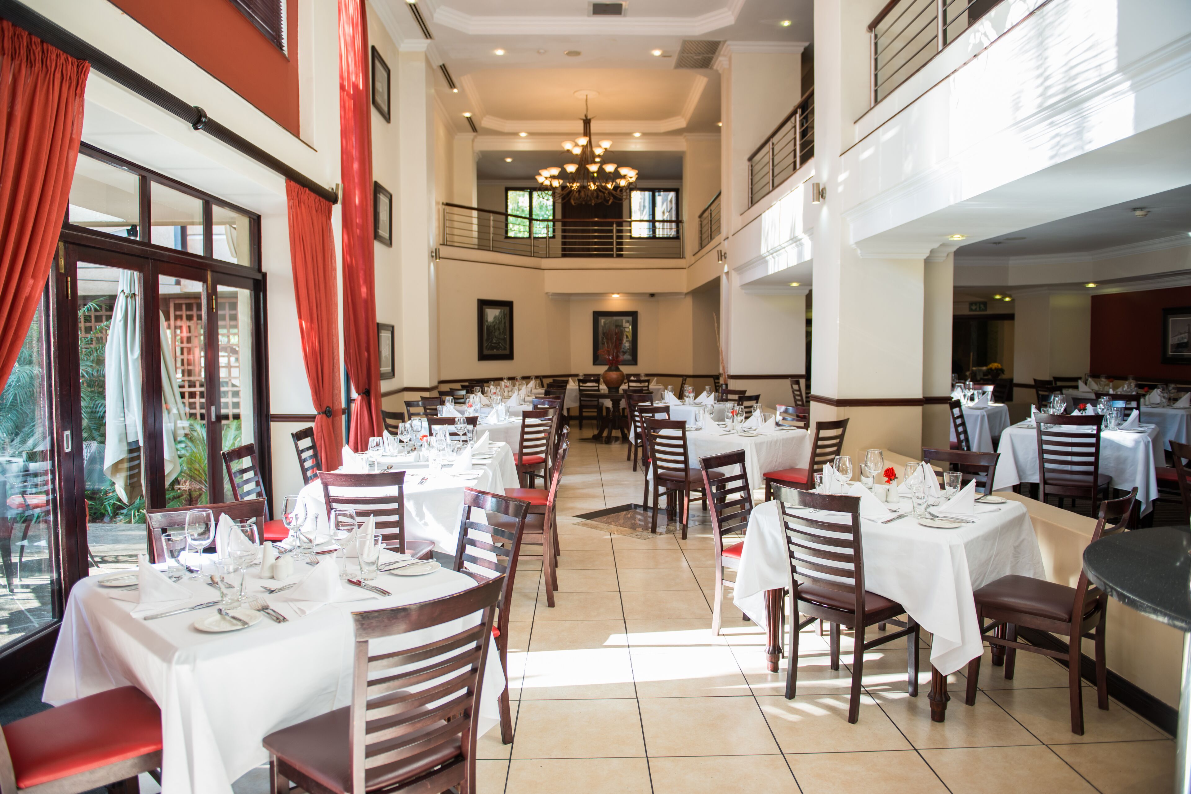 A spacious dining area with multiple tables set for dining, large windows with red curtains, and a chandelier.