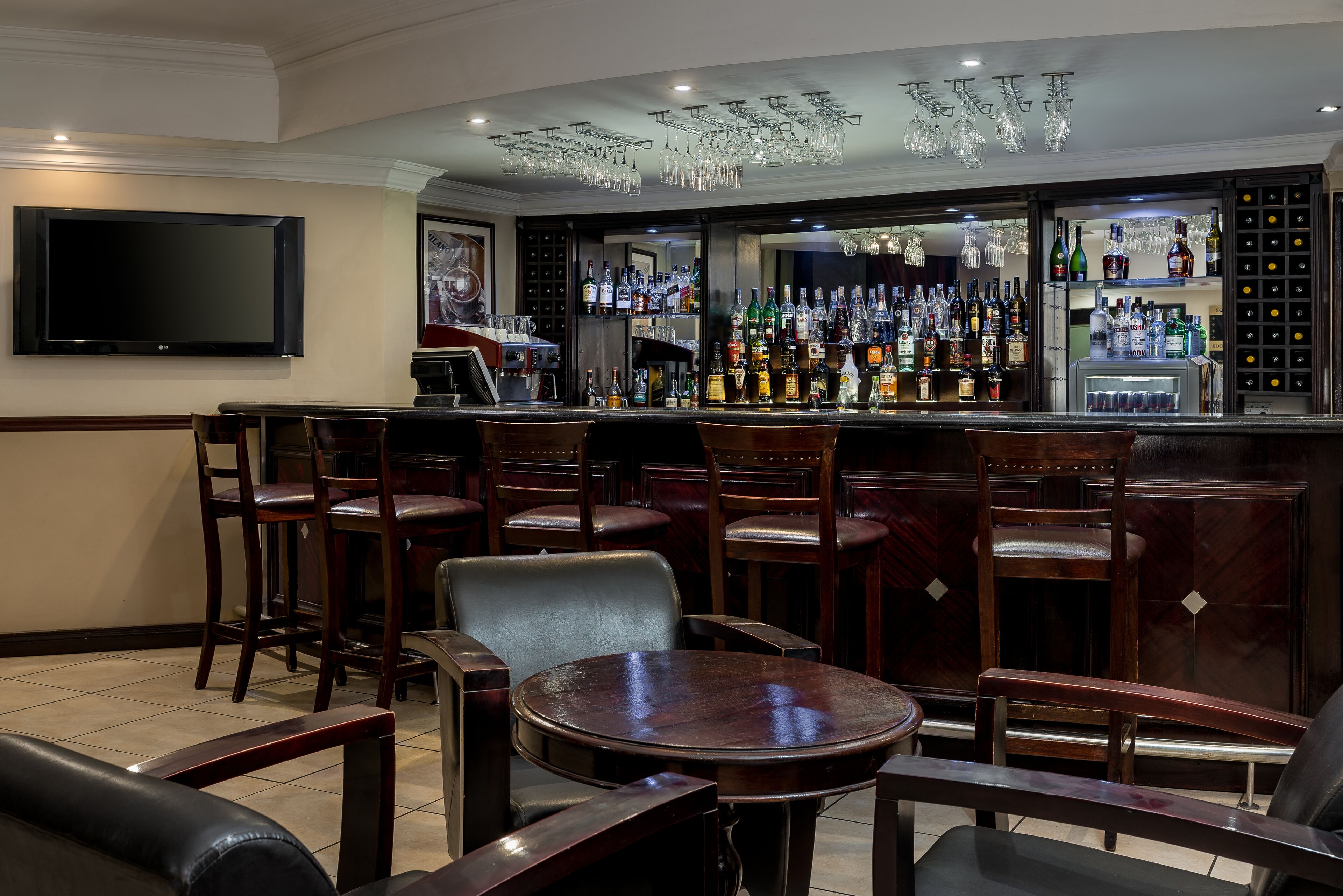 Een bar met diverse zitmogelijkheden, een televisie aan de muur en een assortiment flessen en glazen op de planken.