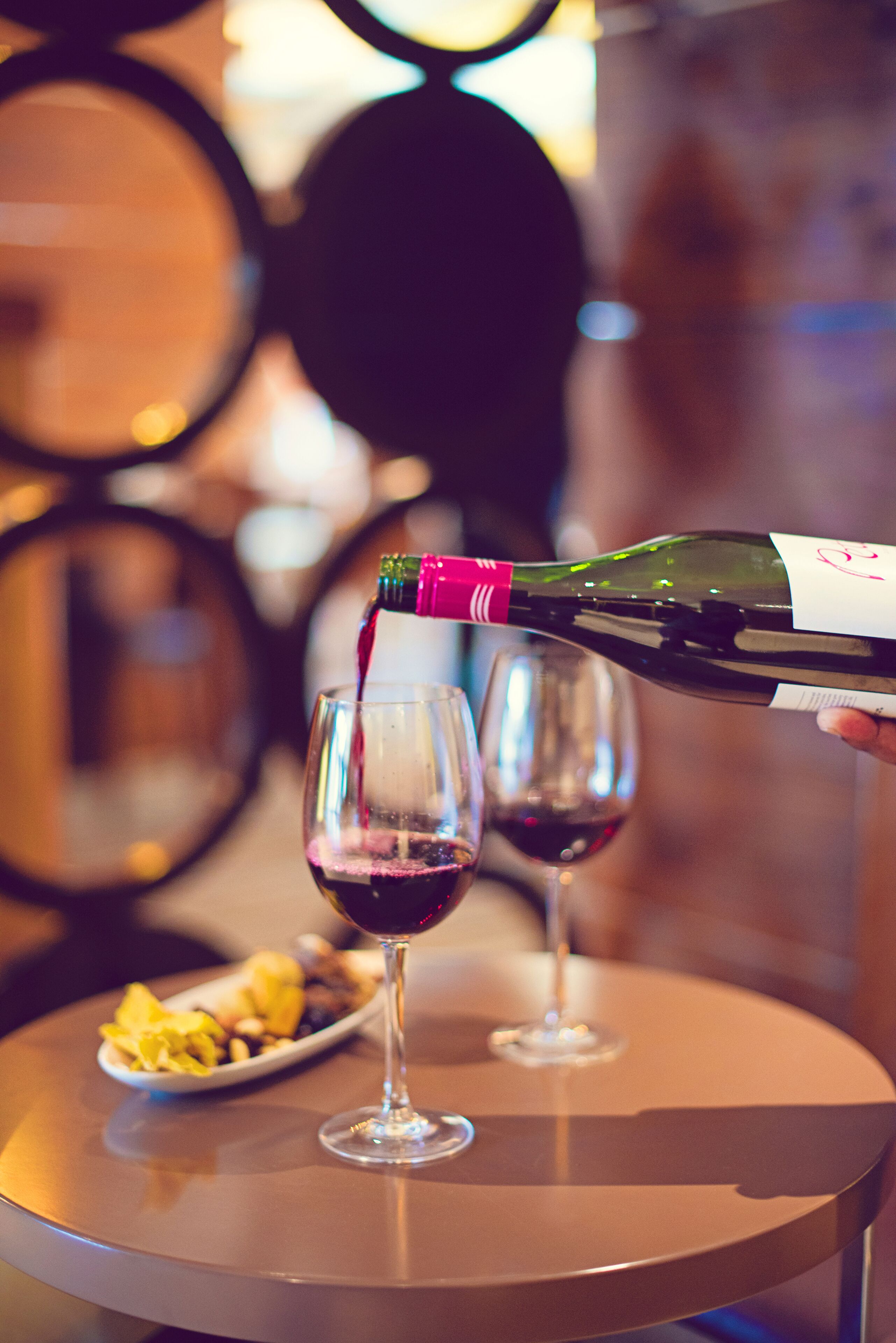 A bottle of wine being poured into a glass on a table with another glass and a plate of food in the background.