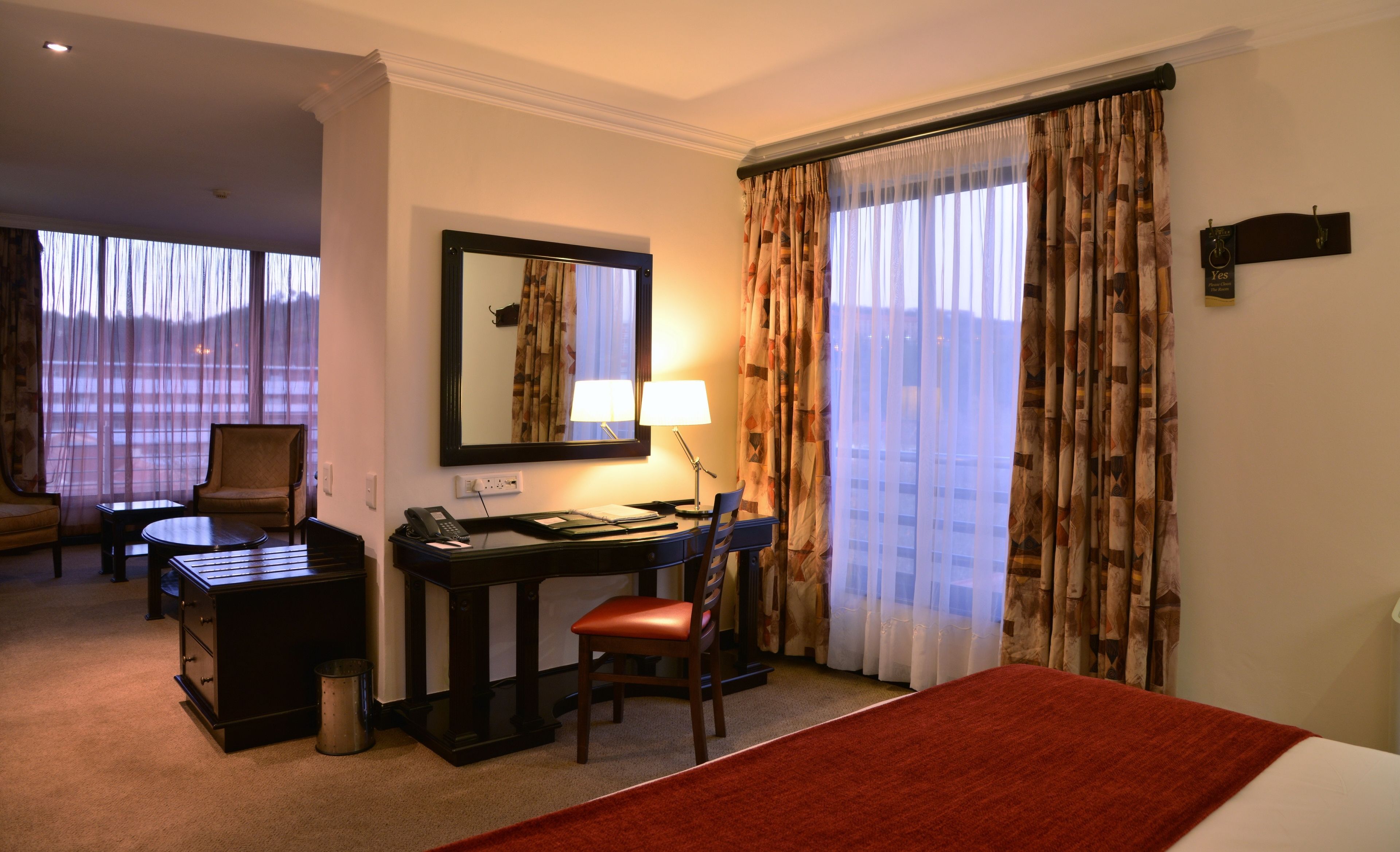 Ein Hotelzimmer mit Schreibtisch, Stuhl und Bett. An der Wand hängt ein Spiegel, auf dem Schreibtisch stehen eine Lampe und ein Telefon. Das Zimmer verfügt über Vorhänge und einen Ausblick nach draußen.