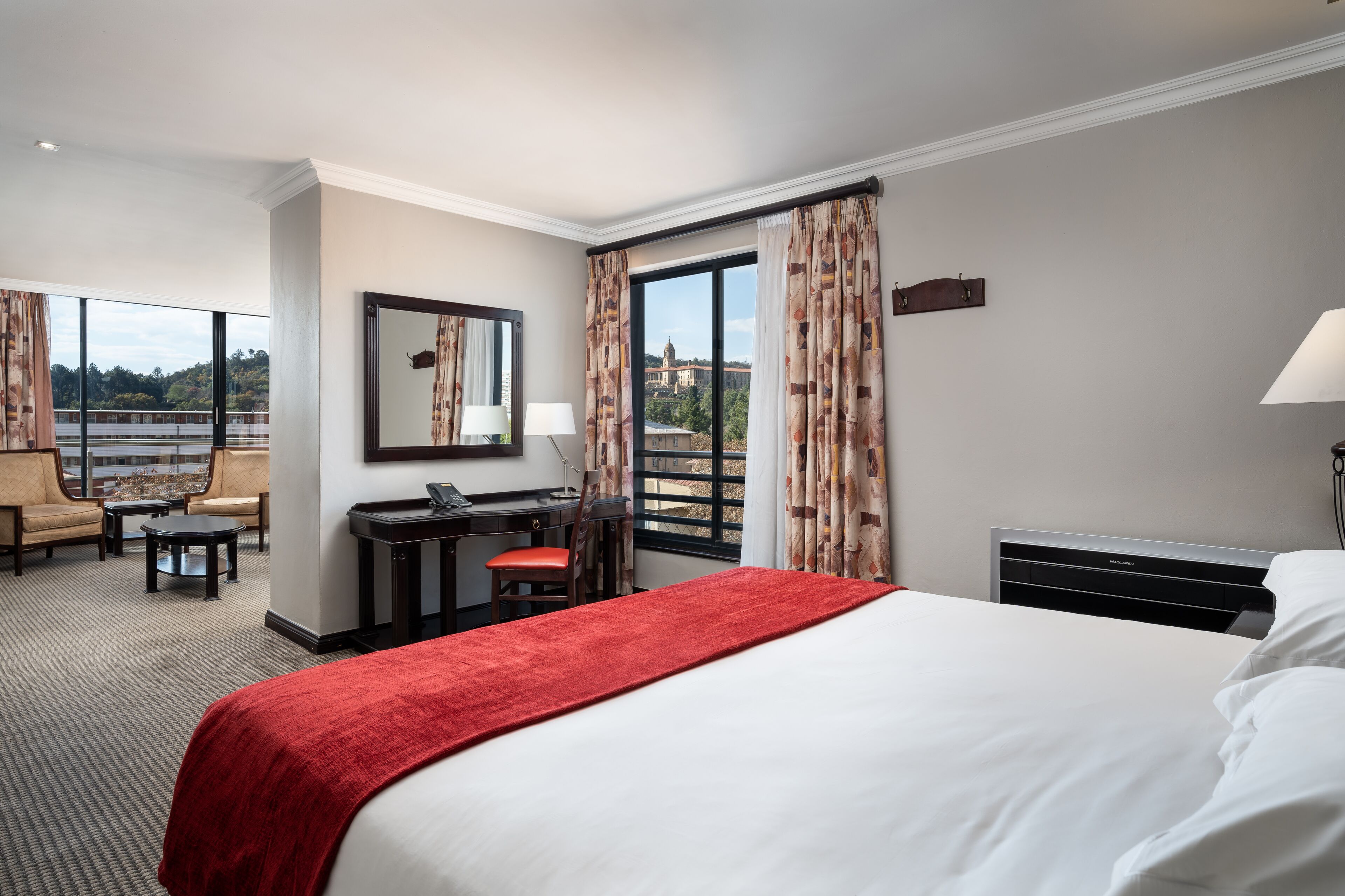 A hotel room with a large bed, a desk, a chair, and a view of a cityscape through large windows.