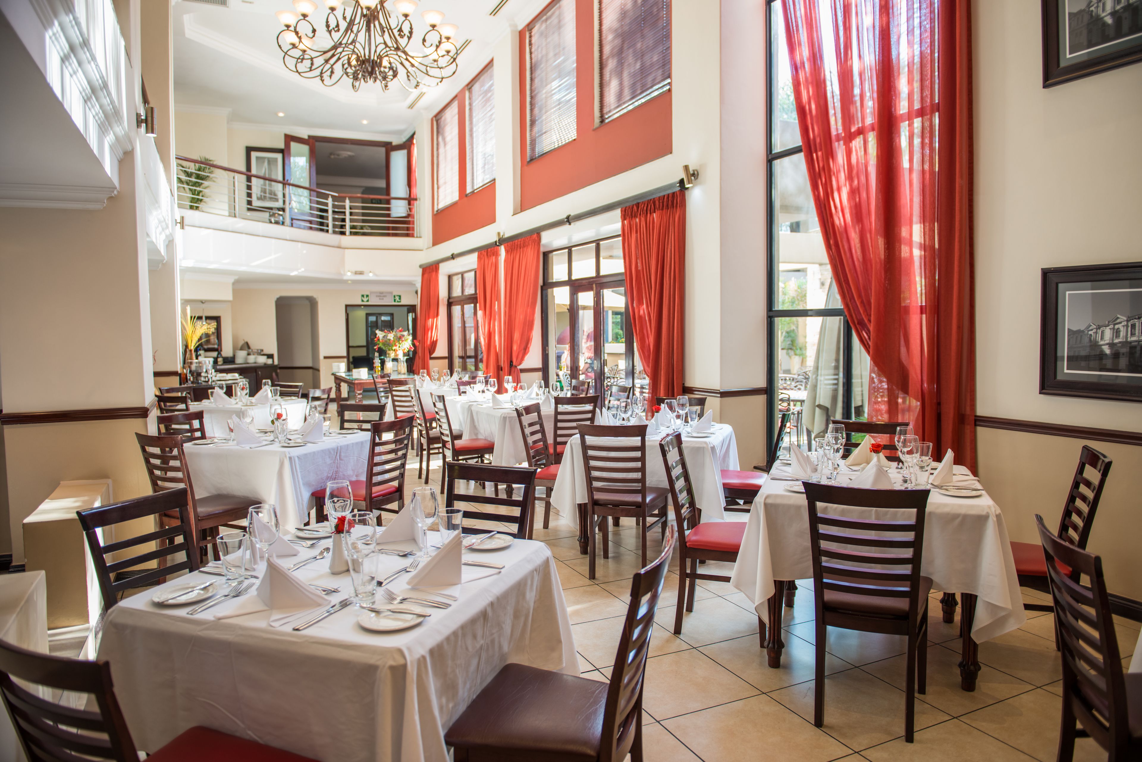 Une salle à manger avec plusieurs tables dressées, de grandes fenêtres ornées de rideaux rouges et un lustre.