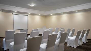 Un salón de conferencias con hileras de sillas cubiertas con fundas blancas, una pantalla de proyector y una mesa.