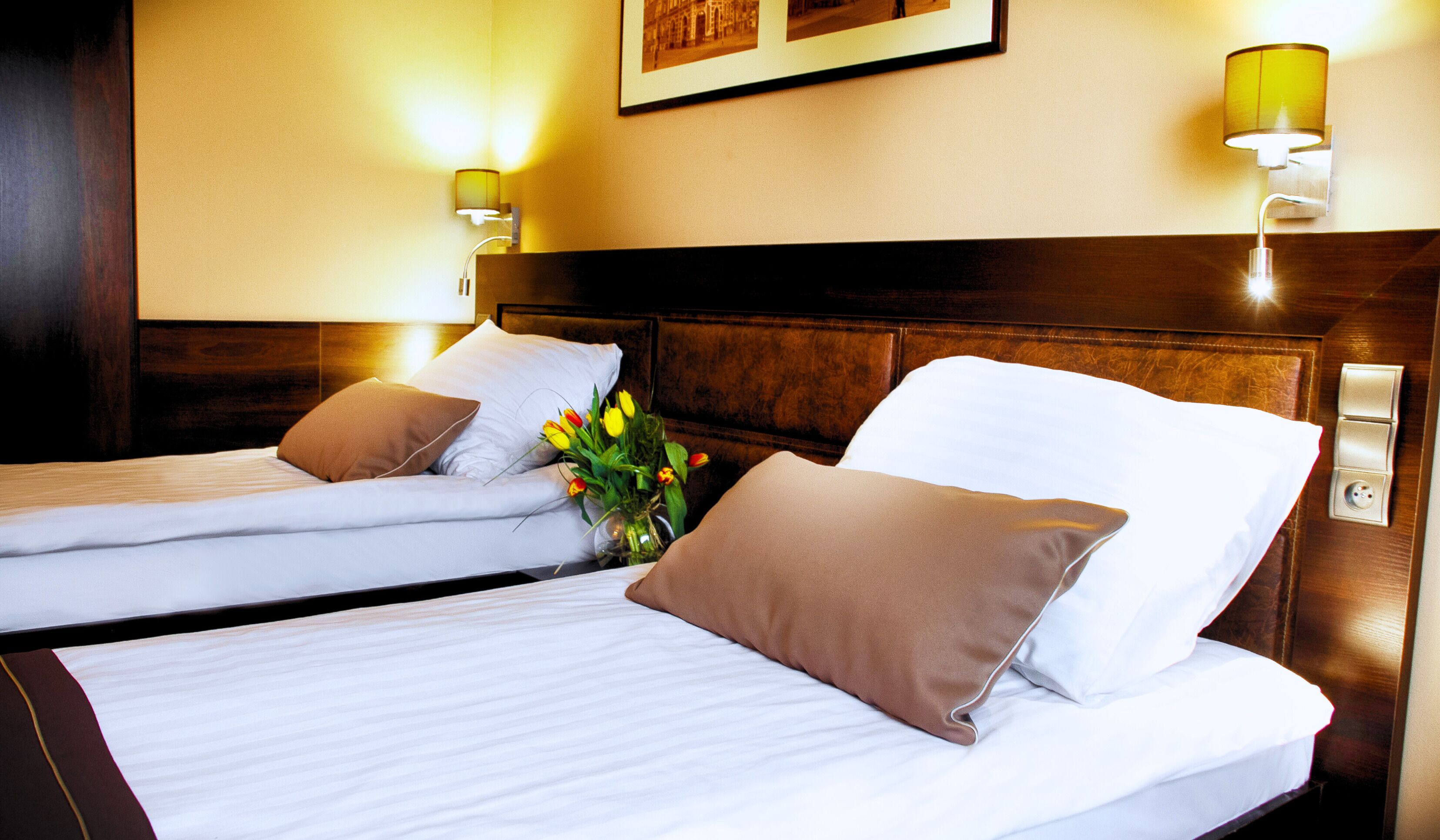 A hotel room with two beds, a wooden headboard, a vase with flowers, and wall-mounted lamps.