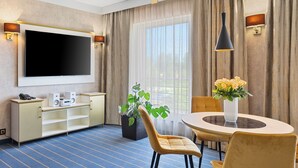 Habitación de hotel con una televisión de pantalla plana grande, una mesa de comedor con sillas, una planta en maceta y una ventana con cortinas.