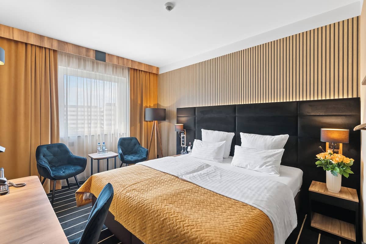 Een hotelkamer met een groot bed, twee stoelen, een bureau met telefoon en een nachtkastje met lamp en bloemen.