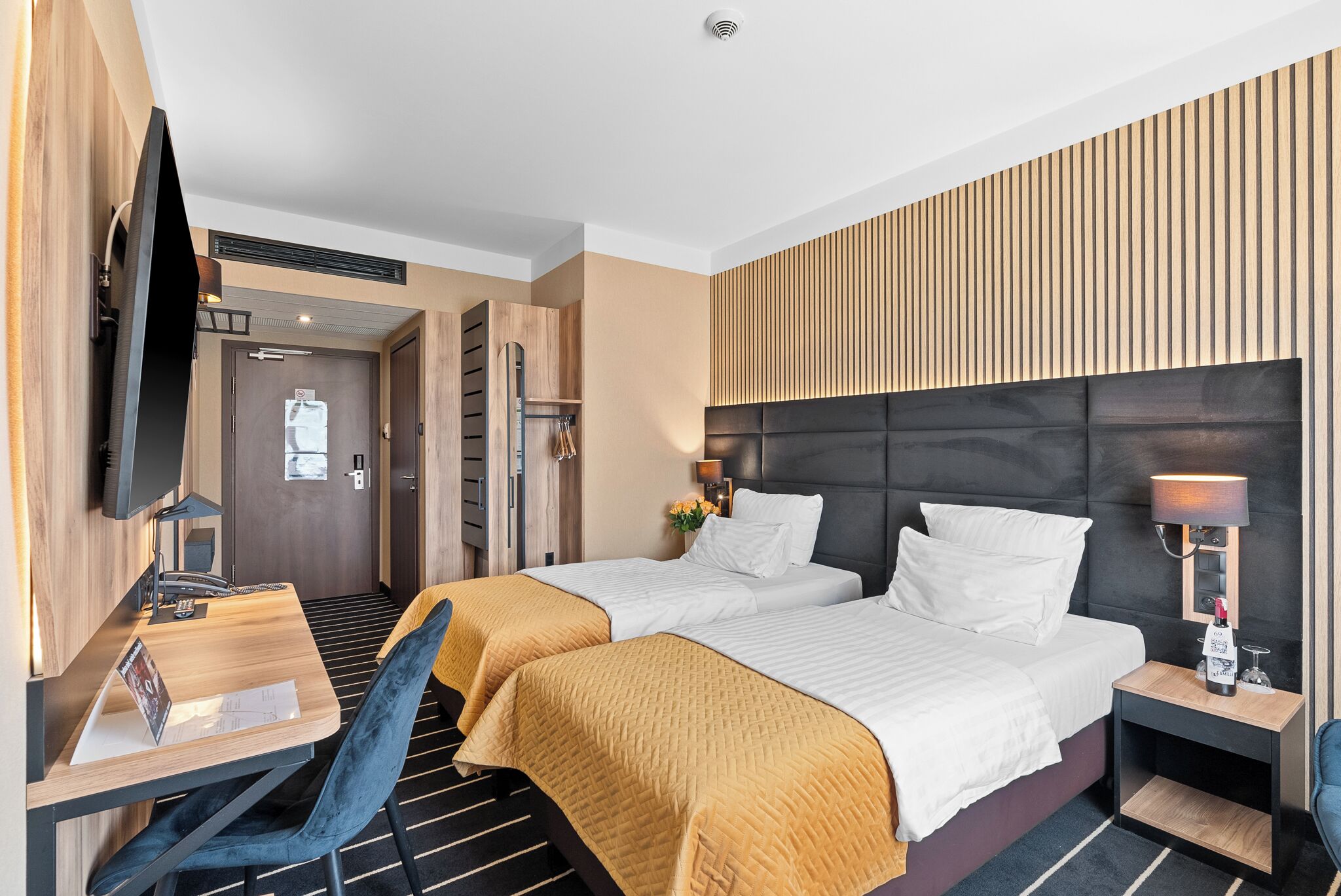 A hotel room with two beds, a desk, a chair, a TV, and a bedside table with a lamp.