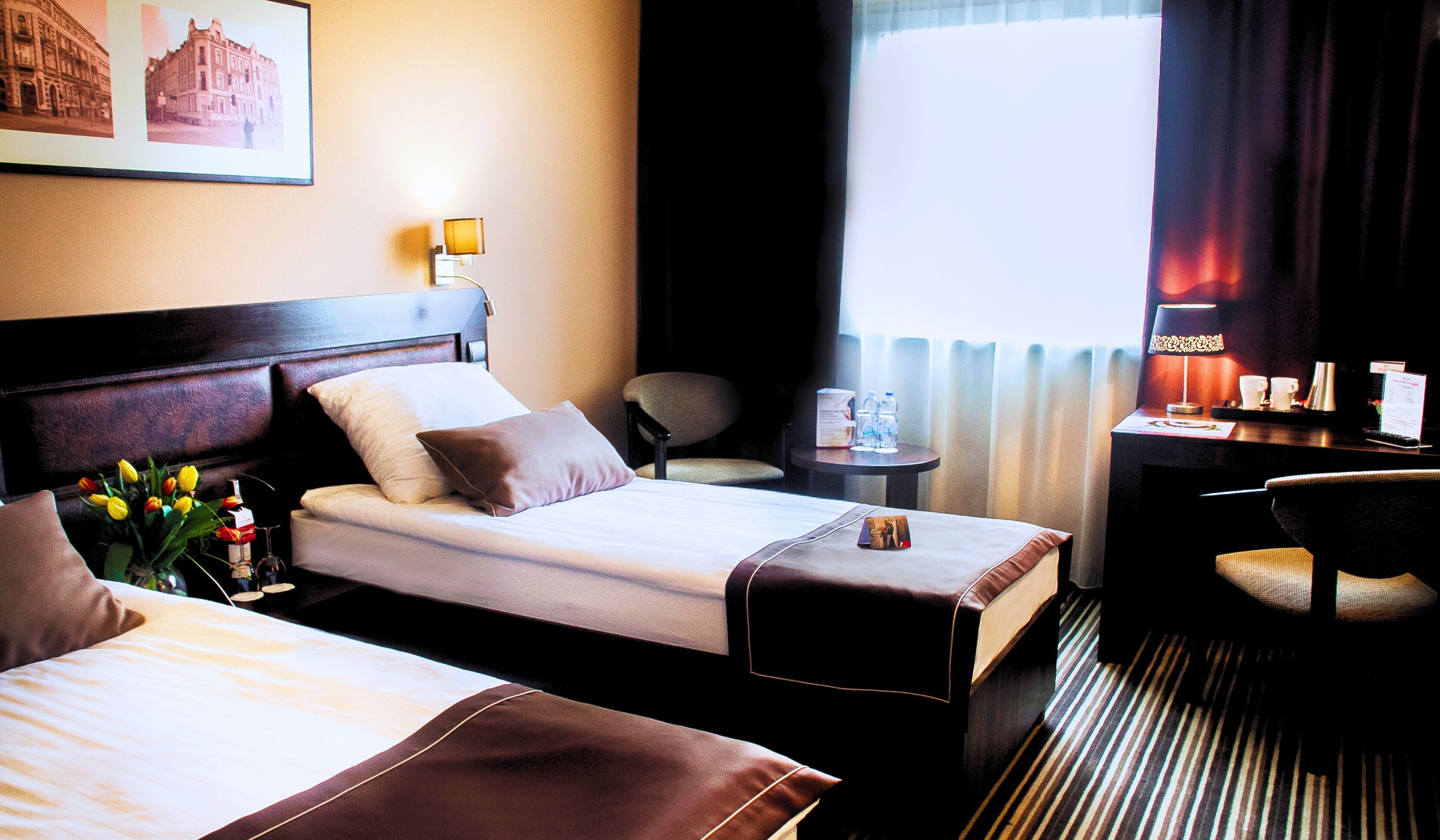 A hotel room with two beds, a desk, a chair, a small table with water bottles, and a lamp.