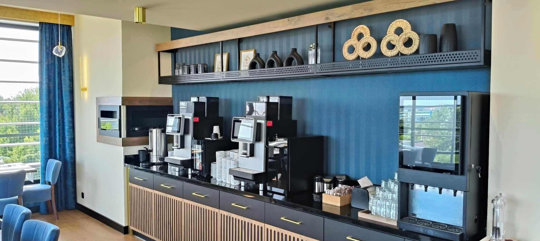 Un bar de hotel con varias máquinas de café, una zona de estar con sillas y vistas a zonas verdes.