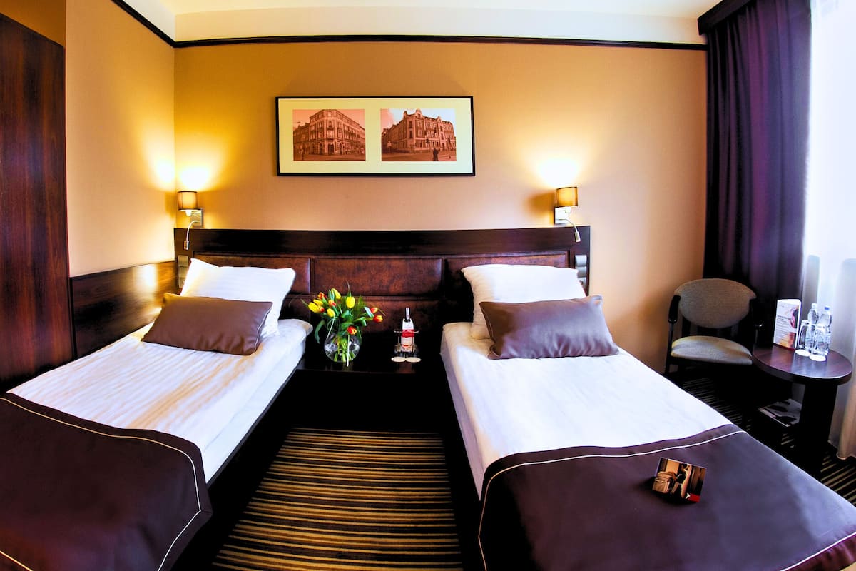 Une chambre d’hôtel avec deux lits, une table de chevet ornée d’un vase de fleurs, un tableau encadré au mur et une chaise près de la fenêtre.
