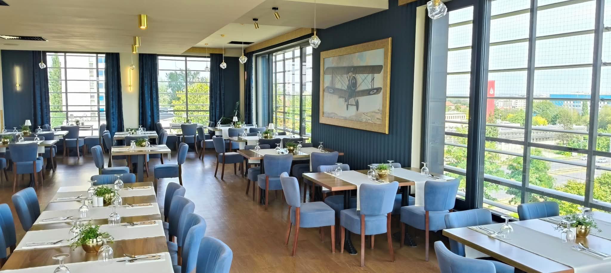 A spacious dining area with multiple tables and chairs, large windows offering a view of the city, and a painting on the wall.