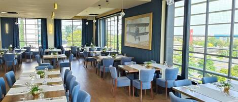 A spacious dining area with multiple tables and chairs, large windows offering a view of the city, and a painting on the wall.